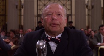 Movie still from “Quiz Show” (1994), directed by Robert Redford – An older man wearing a suit and bow tie is sitting in front of a microphone in front of a crowd; Close Up shot, Low angle