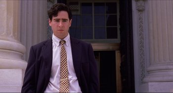 Movie still from “Quiz Show” (1994), directed by Robert Redford – A man in a suit and tie standing in front of a building; Medium shot, Low angle