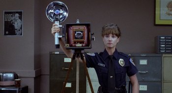 Movie still from “Raising Arizona” (1987), directed by Joel Coen – A police officer holding a camera on top of a tripod; Medium shot, Low angle
