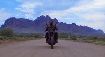 Movie still from “Raising Arizona” (1987), directed by Joel Coen – A man riding a motorcycle down the middle of a dirt road; Wide shot, Low angle