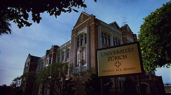 Movie still from “Re-Animator” (1985), directed by Stuart Gordon – A building with a sign in front of it that reads " universität zürich "; Extreme Wide shot, Low angle