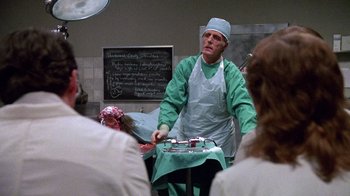 Movie still from “Re-Animator” (1985), directed by Stuart Gordon – A man in a surgeon's outfit stands in front of a group of people; Medium shot, Over the shoulder angle