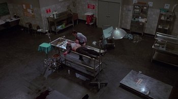 Movie still from “Re-Animator” (1985), directed by Stuart Gordon – A man is laying on a hospital bed in a room; Wide shot, High angle