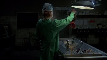 Movie still from “Re-Animator” (1985), directed by Stuart Gordon – A person in a surgical gown and a surgical cap; Medium shot, Over the shoulder angle
