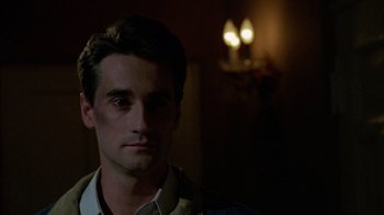 Movie still from “Re-Animator” (1985), directed by Stuart Gordon – A young man with dark hair and a white shirt is looking at the camera; Close Up shot, Over the shoulder angle