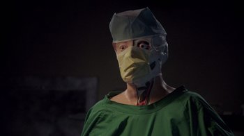 Movie still from “Re-Animator” (1985), directed by Stuart Gordon – A man wearing a surgical mask and a green shirt; Close Up shot, High angle