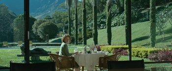 Movie still from “Reaching for the Moon” (2013), directed by Bruno Barreto – A woman sitting at an outdoor table in a garden; Extreme Wide shot, Over the shoulder angle