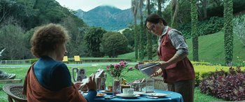 Movie still from “Reaching for the Moon” (2013), directed by Bruno Barreto – A woman standing at a table with a plate of food in front of her; Wide shot, Over the shoulder angle