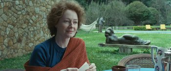 Movie still from “Reaching for the Moon” (2013), directed by Bruno Barreto – A woman sitting at a table in a yard; Medium shot, Over the shoulder angle