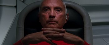 Movie still from “Red Planet” (2000), directed by Antony Hoffman – An older man wearing a red t - shirt is holding his hands near his face; Close Up shot, Overhead angle