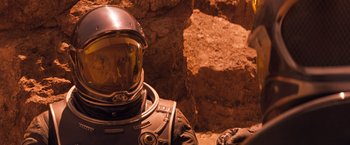 Movie still from “Red Planet” (2000), directed by Antony Hoffman – A person in a space suit is sitting in front of a rock wall; Close Up shot, Over the shoulder angle