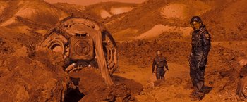 Movie still from “Red Planet” (2000), directed by Antony Hoffman – A man standing next to a machine in a dirt field; Wide shot, High angle