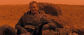 Movie still from “Red Planet” (2000), directed by Antony Hoffman – A man in a space suit sitting next to some rocks; Medium shot, High angle
