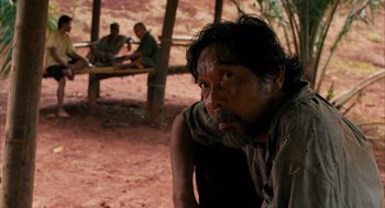 Movie still from “Rescue Dawn” (2006), directed by Werner Herzog – A man sitting on the ground in front of a wooden bench; Medium shot, Over the shoulder angle