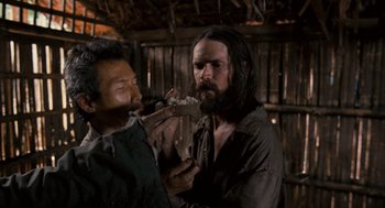 Movie still from “Rescue Dawn” (2006), directed by Werner Herzog – A man with a beard is eating something while another man looks on; Close Up shot, Over the shoulder angle