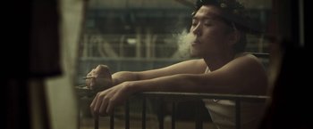 Movie still from “Revenge of the Green Dragons” (2014), directed by Andrew Loo – A man sitting on a railing smoking a cigarette; Medium shot, Low angle