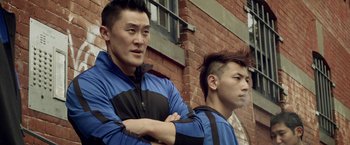 Movie still from “Revenge of the Green Dragons” (2014), directed by Andrew Loo – Two asian men standing next to each other in front of a brick building; Close Up shot, Low angle