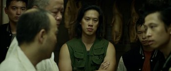 Movie still from “Revenge of the Green Dragons” (2014), directed by Andrew Loo – A man with long hair and a green vest is standing with two other men; Medium shot, Over the shoulder angle