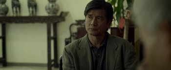 Movie still from “Revenge of the Green Dragons” (2014), directed by Andrew Loo – A man sitting in a chair in front of a plant; Close Up shot, Over the shoulder angle