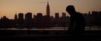 Movie still from “Revenge of the Green Dragons” (2014), directed by Andrew Loo – A man sitting on a bench in front of a city skyline; Wide shot, Low angle