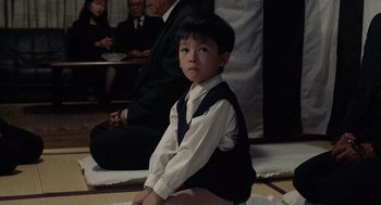 Movie still from “Ringu” (1998), directed by Hideo Nakata – A young boy sitting on the floor in front of a group of people; Medium shot, High angle