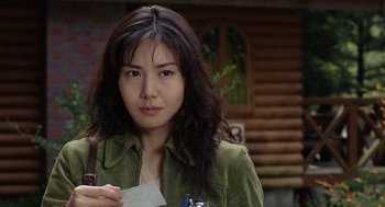 Movie still from “Ringu” (1998), directed by Hideo Nakata – A woman holding a business card while standing in front of a log cabin; Close Up shot, Over the shoulder angle
