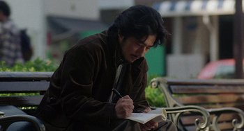 Movie still from “Ringu” (1998), directed by Hideo Nakata – A man sitting on a park bench writing on a book; Medium shot, Low angle