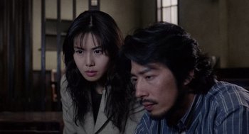 Movie still from “Ringu” (1998), directed by Hideo Nakata – A woman standing next to a man sitting next to a window; Close Up shot, High angle