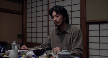 Movie still from “Ringu” (1998), directed by Hideo Nakata – A man sitting at a table with a bowl of food; Medium shot, Low angle