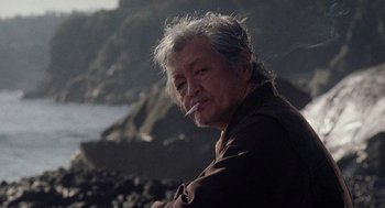 Movie still from “Ringu” (1998), directed by Hideo Nakata – An older man sitting on the beach smoking a cigarette; Close Up shot, Low angle