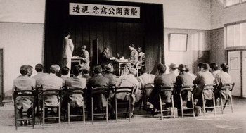 Movie still from “Ringu” (1998), directed by Hideo Nakata – A group of people sitting in chairs in front of a stage; Wide shot, Low angle