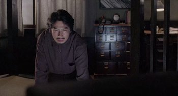 Movie still from “Ringu” (1998), directed by Hideo Nakata – A man sitting on a bed in a dark room; Medium shot, Over the shoulder angle