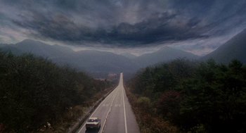 Movie still from “Ringu” (1998), directed by Hideo Nakata – A car driving down a road under a cloudy sky; Extreme Wide shot, High angle