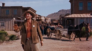 Movie still from “Rio Bravo” (1959), directed by Howard Hawks – A man wearing a cowboy hat and a brown jacket walking down a street; Medium shot, Low angle