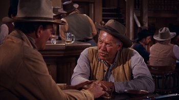 Movie still from “Rio Bravo” (1959), directed by Howard Hawks – Two men sitting at a table in front of other men; Medium shot, Over the shoulder angle