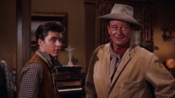 Movie still from “Rio Bravo” (1959), directed by Howard Hawks – A man standing next to another man in a hat; Medium shot, Over the shoulder angle