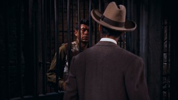 Movie still from “Rio Bravo” (1959), directed by Howard Hawks – A man in a suit and hat standing next to another man in front of a cage; Medium shot, Over the shoulder angle