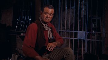 Movie still from “Rio Bravo” (1959), directed by Howard Hawks – A man sitting in front of a metal fence; Medium shot, Low angle