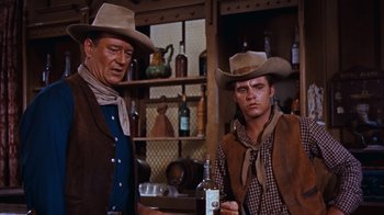 Movie still from “Rio Bravo” (1959), directed by Howard Hawks – Two men in cowboy hats standing next to each other holding a bottle; Medium shot, Low angle