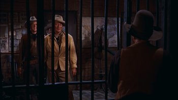 Movie still from “Rio Bravo” (1959), directed by Howard Hawks – A man in a suit and hat standing in front of a fence; Medium shot, Over the shoulder angle