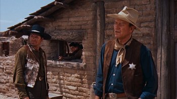 Movie still from “Rio Bravo” (1959), directed by Howard Hawks – A man in a cowboy hat standing in front of a brick building; Medium shot, Over the shoulder angle