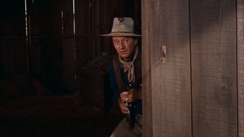 Movie still from “Rio Bravo” (1959), directed by Howard Hawks – A man in a hat holding a cup in a barn; Medium shot, Over the shoulder angle