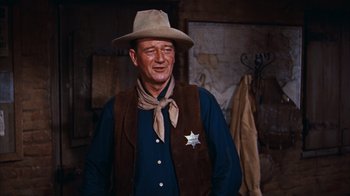 Movie still from “Rio Bravo” (1959), directed by Howard Hawks – A man wearing a cowboy hat and a vest; Medium shot, Over the shoulder angle