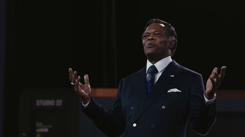 Movie still from “RoboCop” (2014), directed by José Padilha – A man in a suit and tie talking on a stage; Medium shot, Low angle