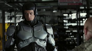 Movie still from “RoboCop” (2014), directed by José Padilha – A man in a suit and helmet standing in a room; Medium shot, Over the shoulder angle
