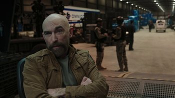 Movie still from “RoboCop” (2014), directed by José Padilha – A bald man in a tan jacket with arms crossed in front of a group of men in fatigues; Medium shot, Over the shoulder angle