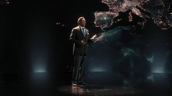 Movie still from “RoboCop” (2014), directed by José Padilha – A man in a suit and tie on a stage; Wide shot, High angle