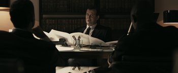 Movie still from “RocknRolla” (2008), directed by Guy Ritchie – A man sitting at a table with papers in front of him; Medium shot, Over the shoulder angle