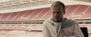 Movie still from “RocknRolla” (2008), directed by Guy Ritchie – A man wearing a white shirt and a gray scarf in front of an empty stadium; Close Up shot, Low angle