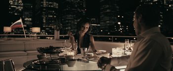 Movie still from “RocknRolla” (2008), directed by Guy Ritchie – A woman sitting at a dinner table in front of a city skyline; Medium shot, Over the shoulder angle